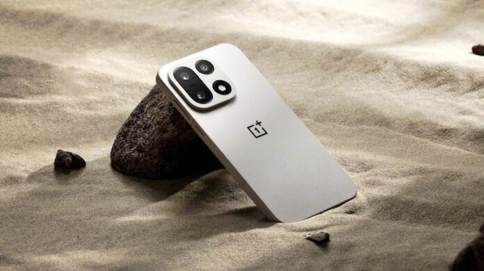 White OnePlus 15 smartphone with triple-camera module, propped against a rock on sand