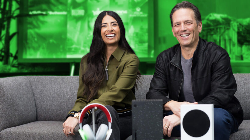 Asha Sharma and another Xbox executive smiling on a couch with Xbox consoles and a headset displayed in front of them
