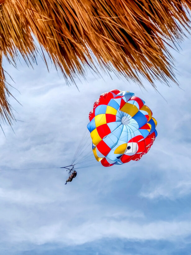 Parasailer with a colorful red, yellow, and blue canopy soaring against a cloudy sky, framed by a thatched palapa roof in the foreground