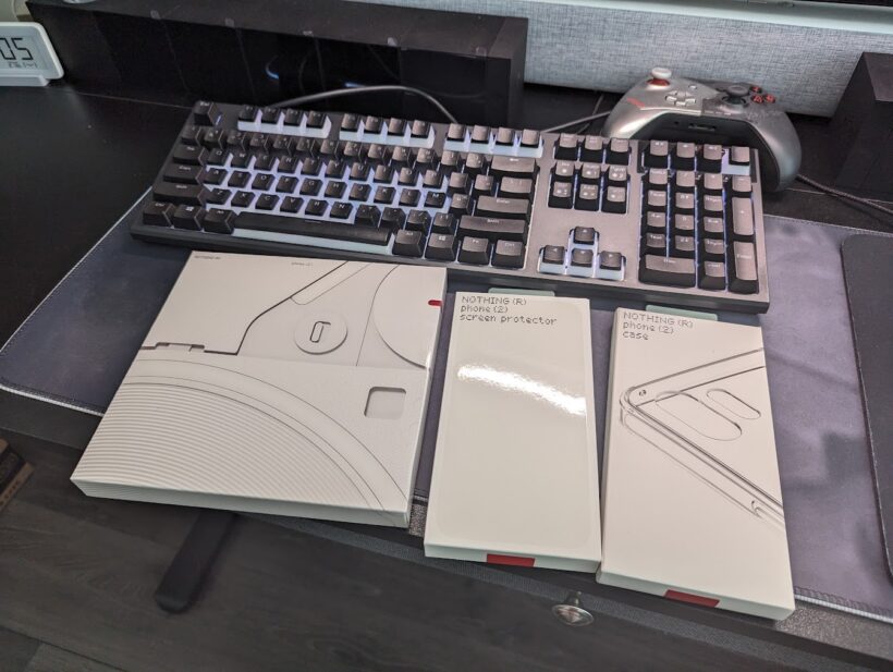 Nothing Phone (2) retail box alongside its official screen protector and case packaging, on a desk with a mechanical keyboard and game controller