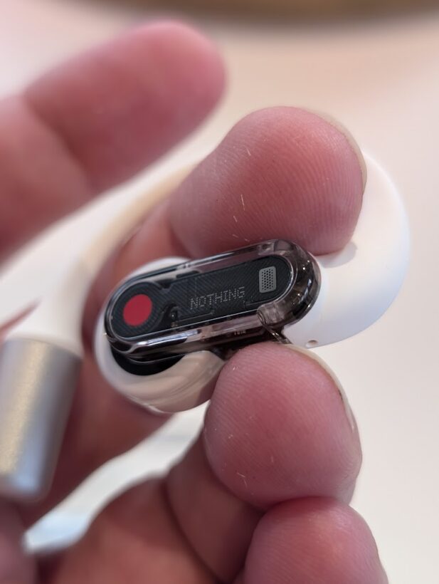 Close-up of a Nothing earbud held between fingers, showing its transparent stem with the Nothing logo and a red driver visible inside