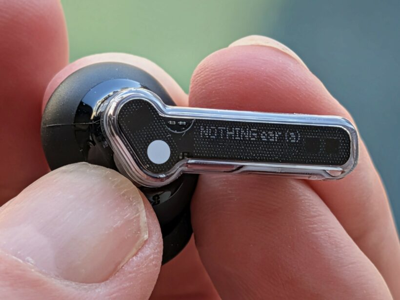 Close-up of a hand holding a Nothing Ear (a) wireless earbud, showing the stem with Nothing branding and transparent design details
