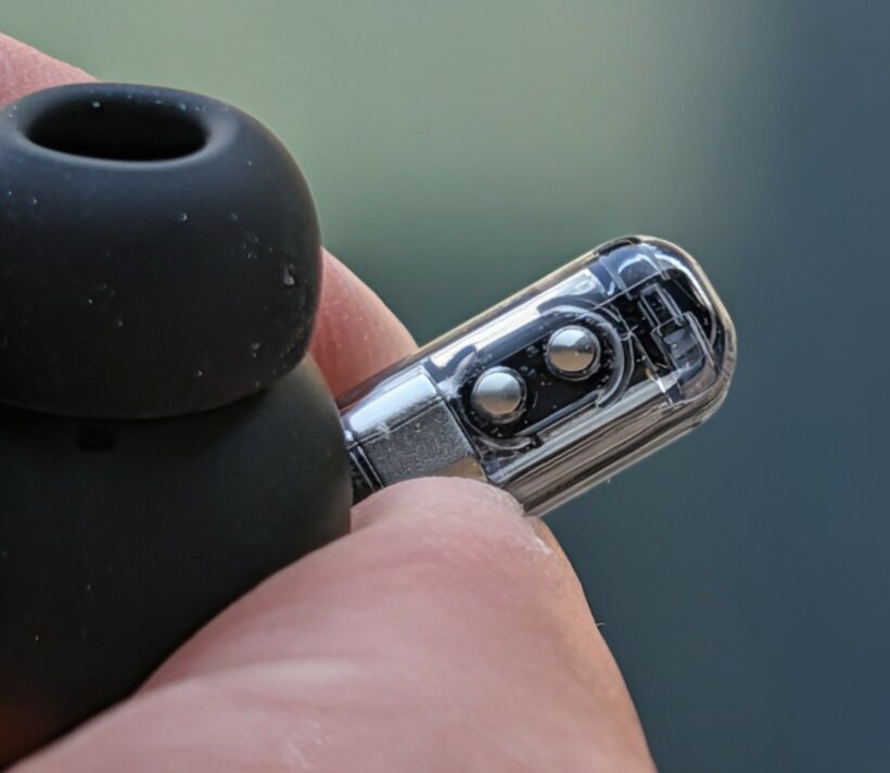 Close-up of a wireless earbud's transparent stem held between fingers, revealing internal sensors and components