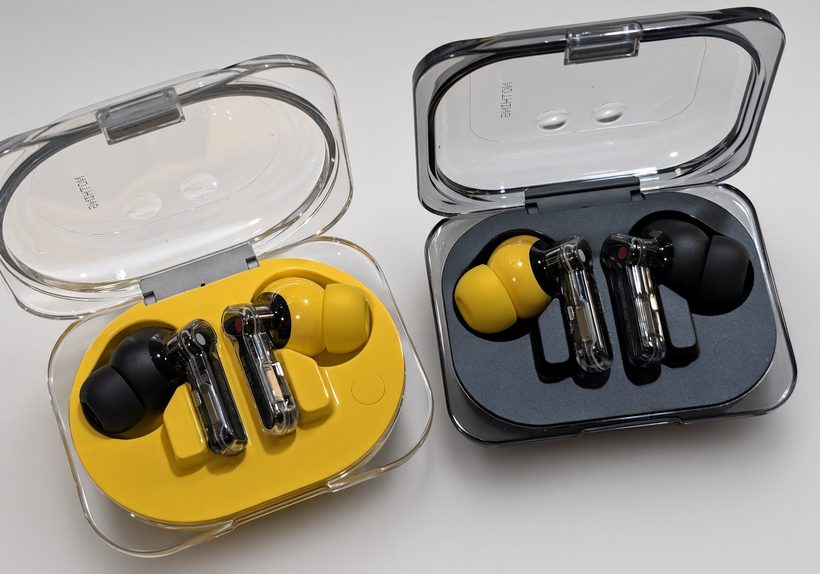 Two open transparent earbud cases side by side, one with a yellow tray and one with a black tray, each holding a pair of clear-bodied earbuds with black and yellow ear tips